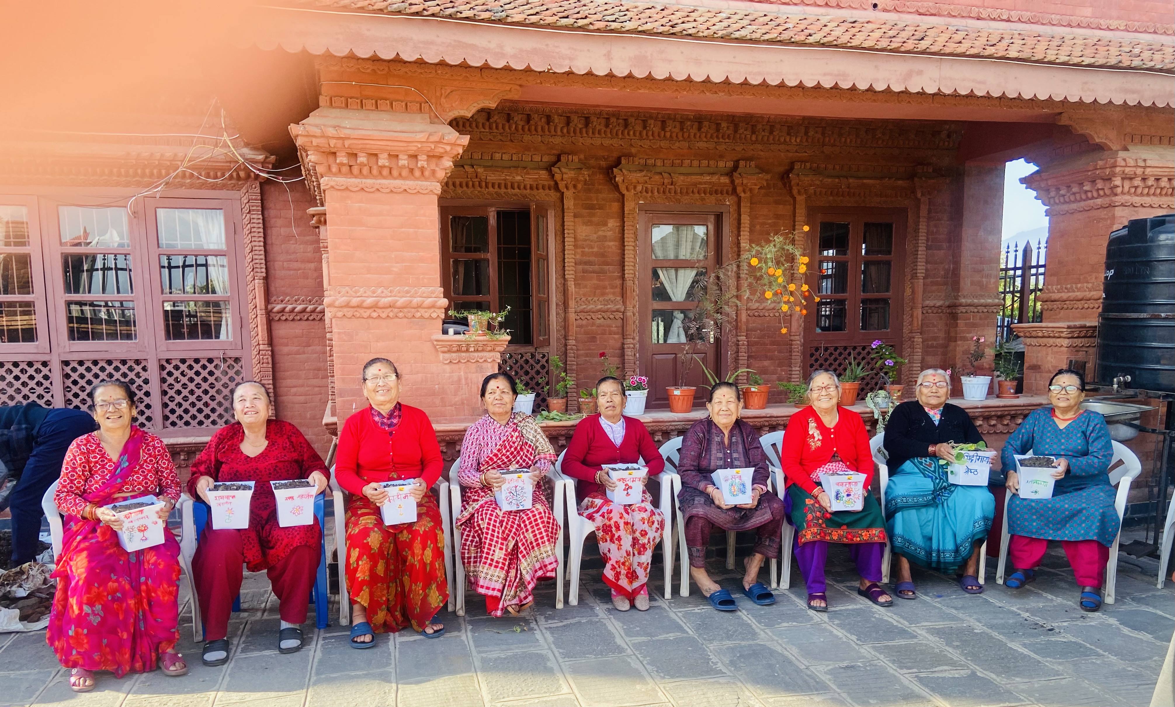 Seniors at Ananda Niketan holding potted plants in front of a traditional Newari brick building
